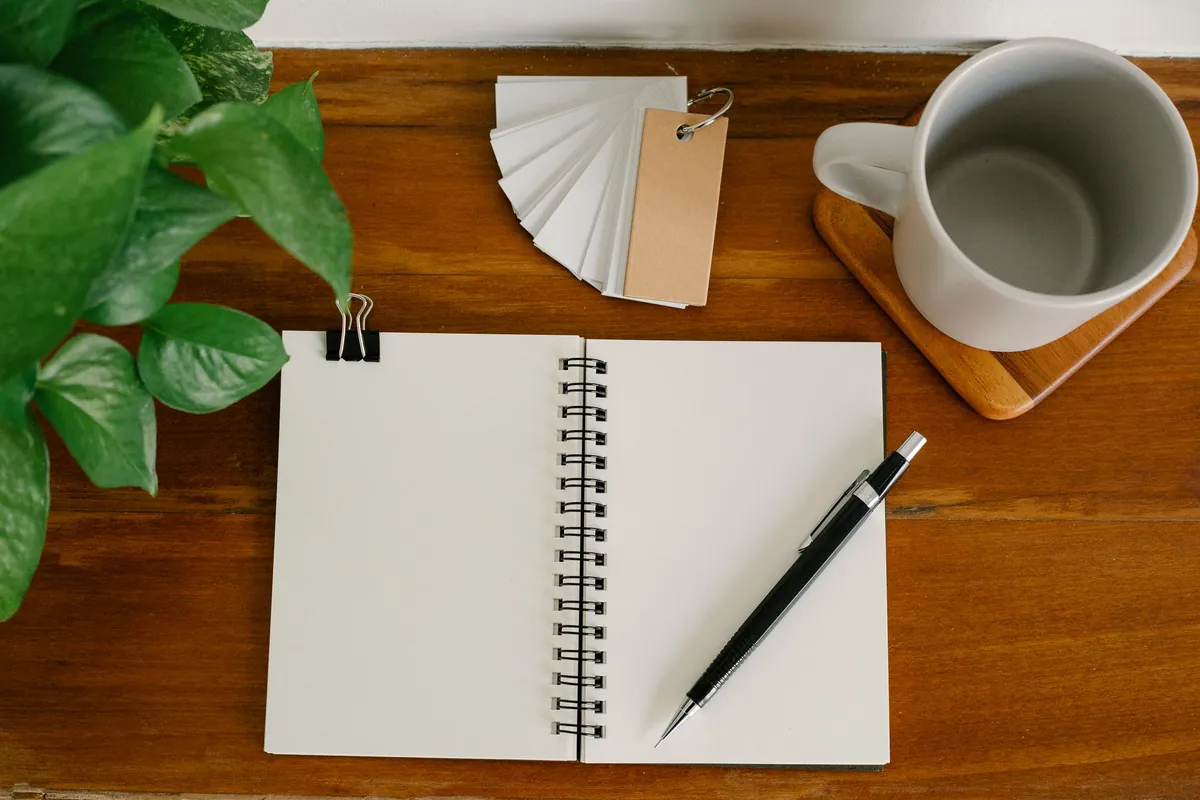An open notebook and a pen on a desk beside a plant and an empty mug.